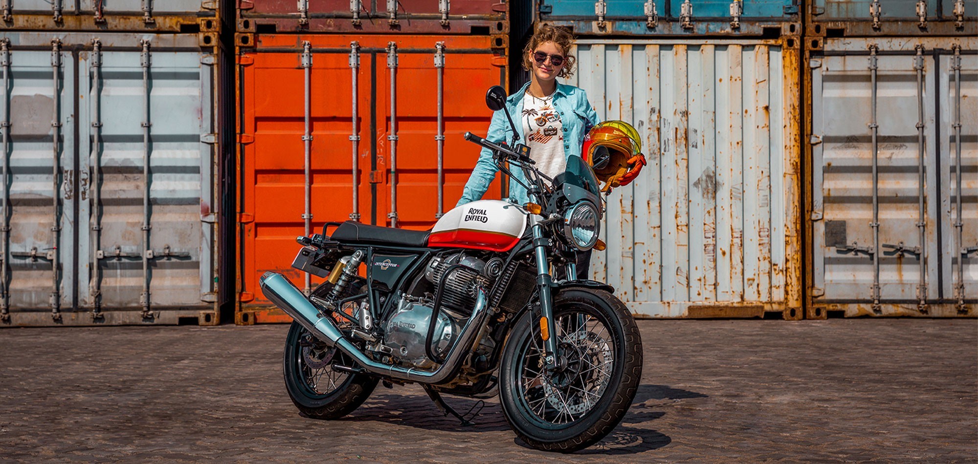 Female Rider standing behind a Royal Enfield Interceptor 650 with colourful shipping containers in the background