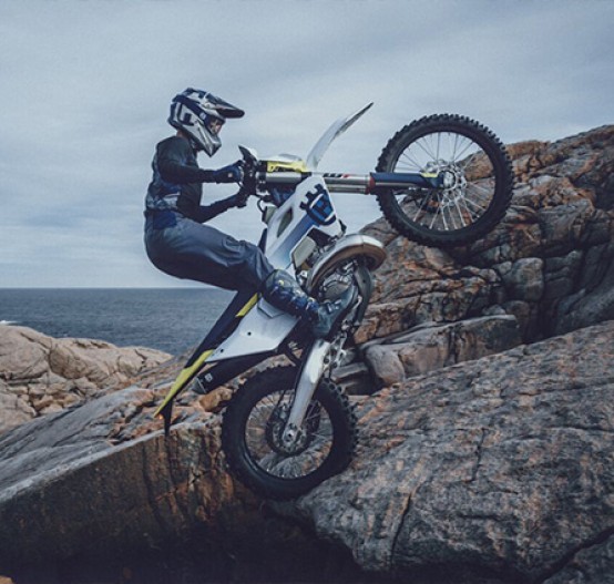 Action image of Husqvarna TE 250/300 in white colourway, wheelie over rocks by the ocean